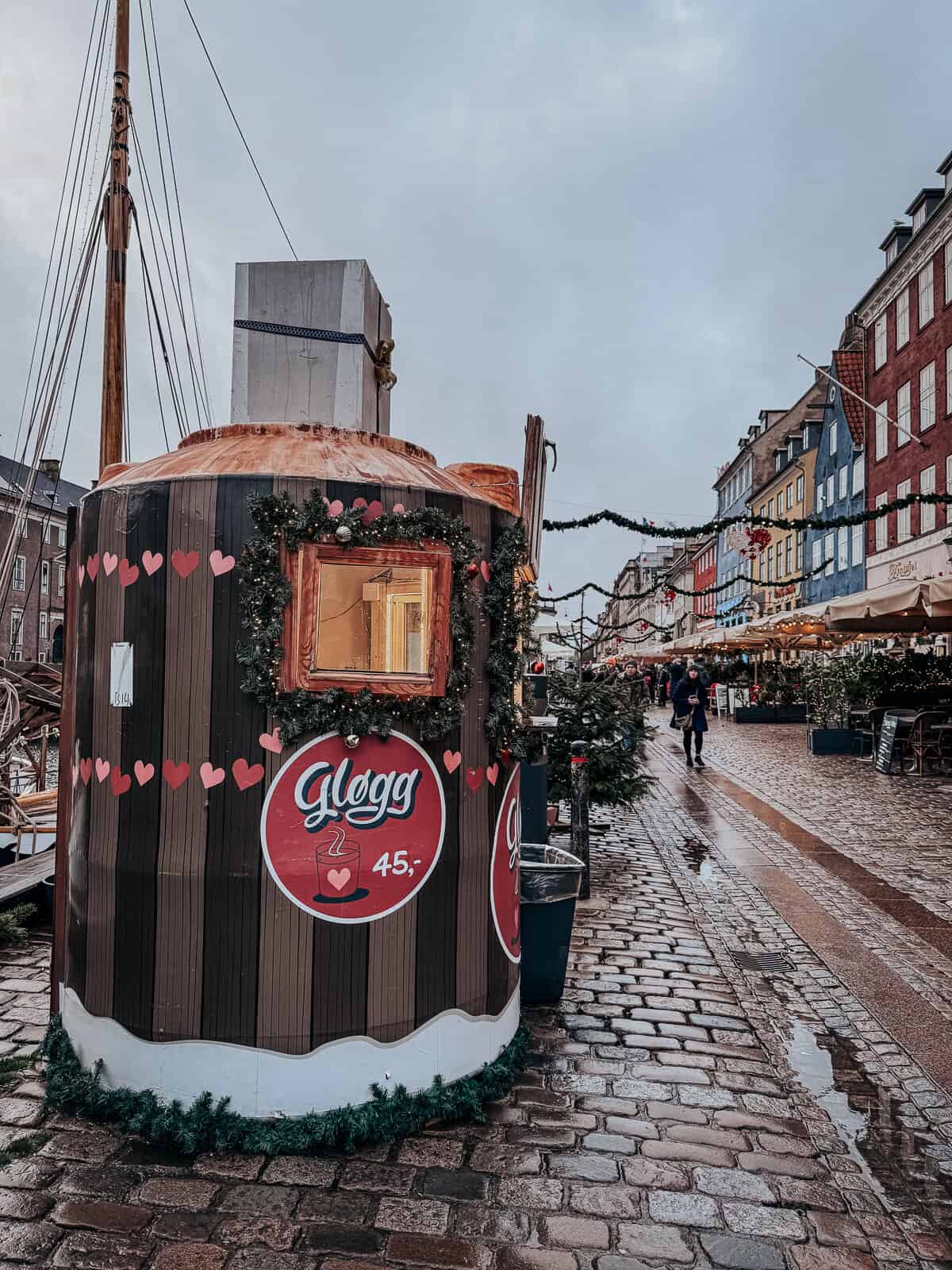 A wooden market stand decorated with Christmas greenery and signs for traditional hot beverages, with customers inside enjoying drinks.