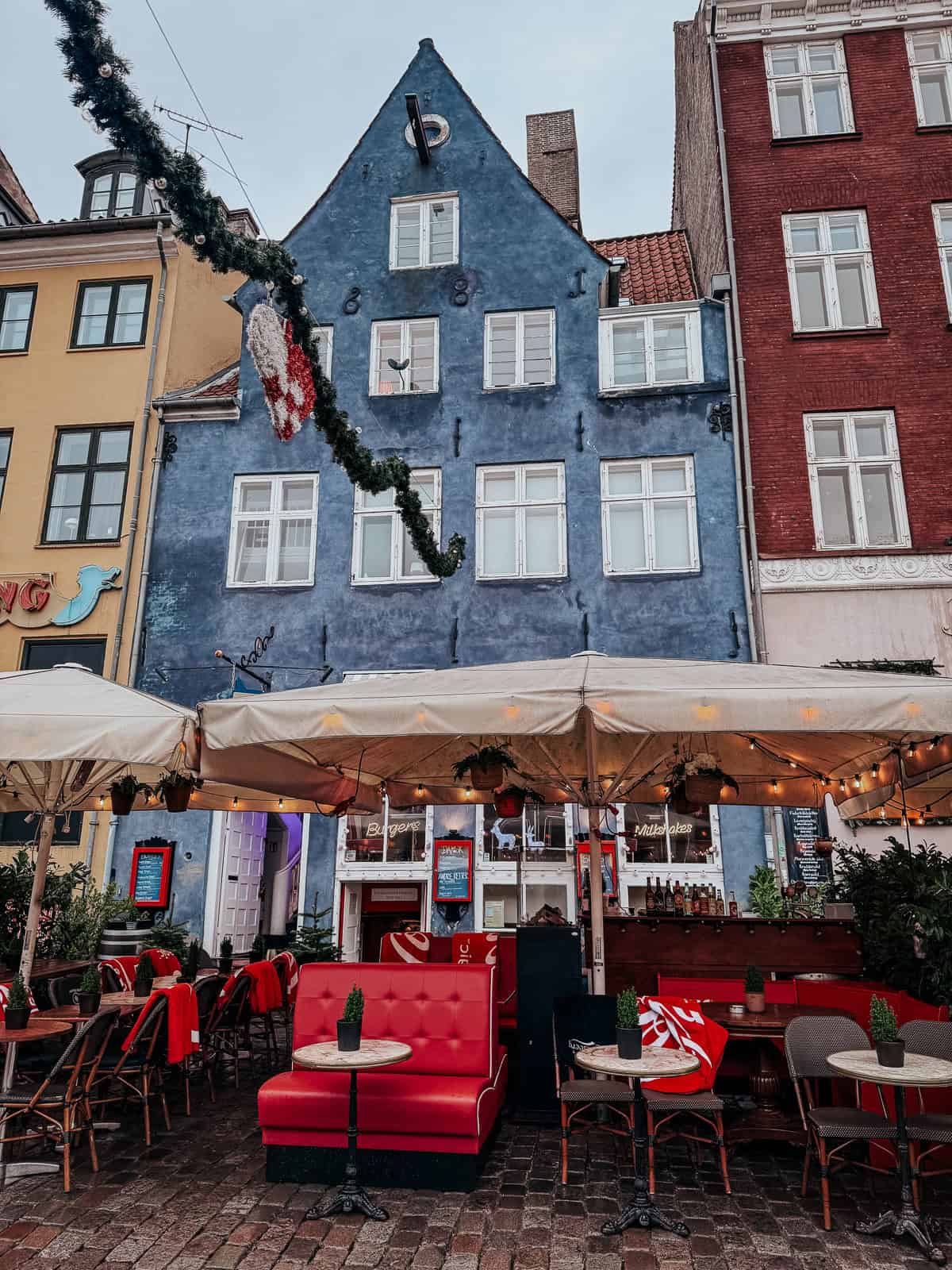 A quaint cafe with outdoor seating, festively decorated for Christmas, set against colorful historic buildings on a cobblestone street.