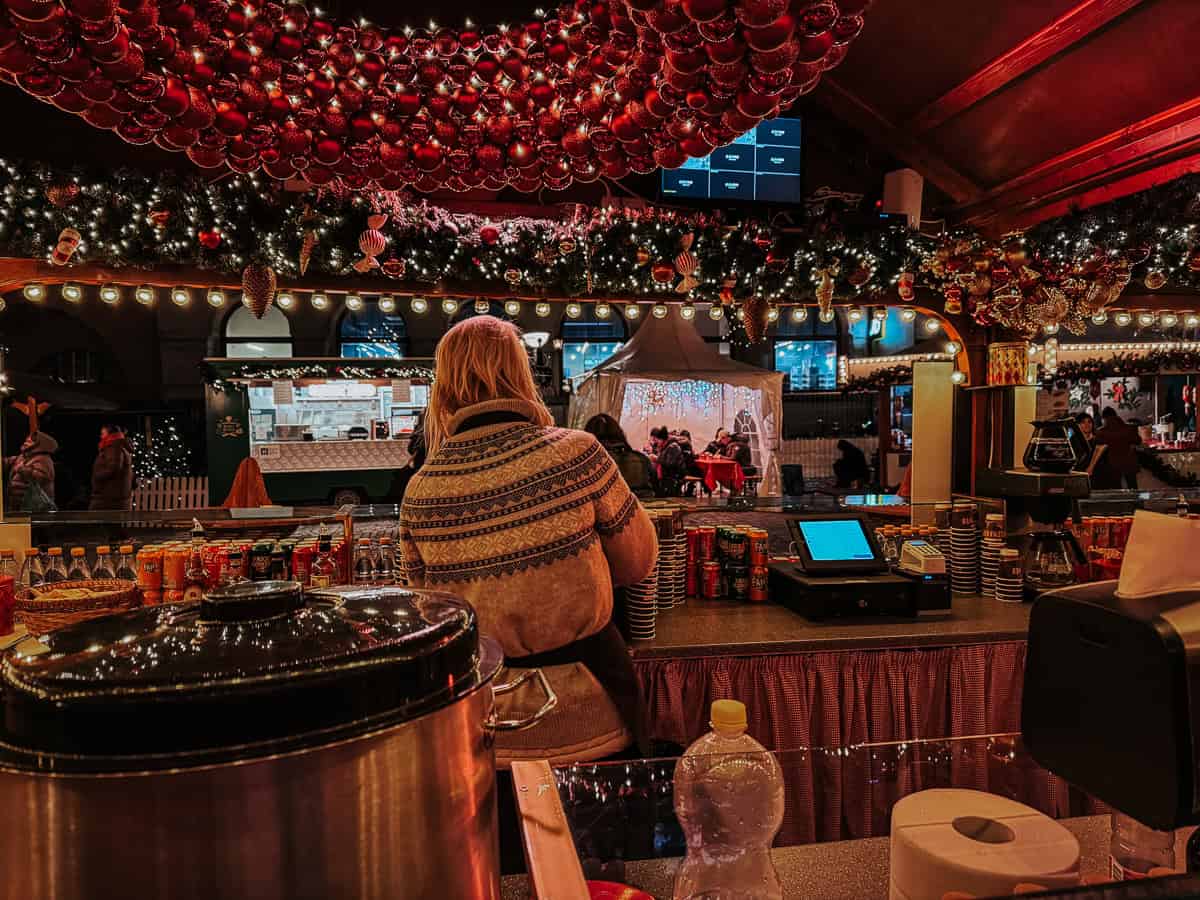 View from a market vendor's stall showing a large pot, beverages, and the back of a vendor looking out at the busy Christmas market decorated with red Christmas balls and lights.
