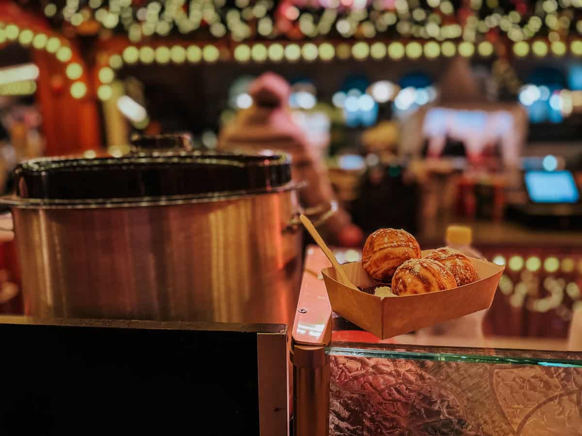 A street food stall at a Christmas market serving pastries in a paper tray, with a large pot and festive lights in the background.