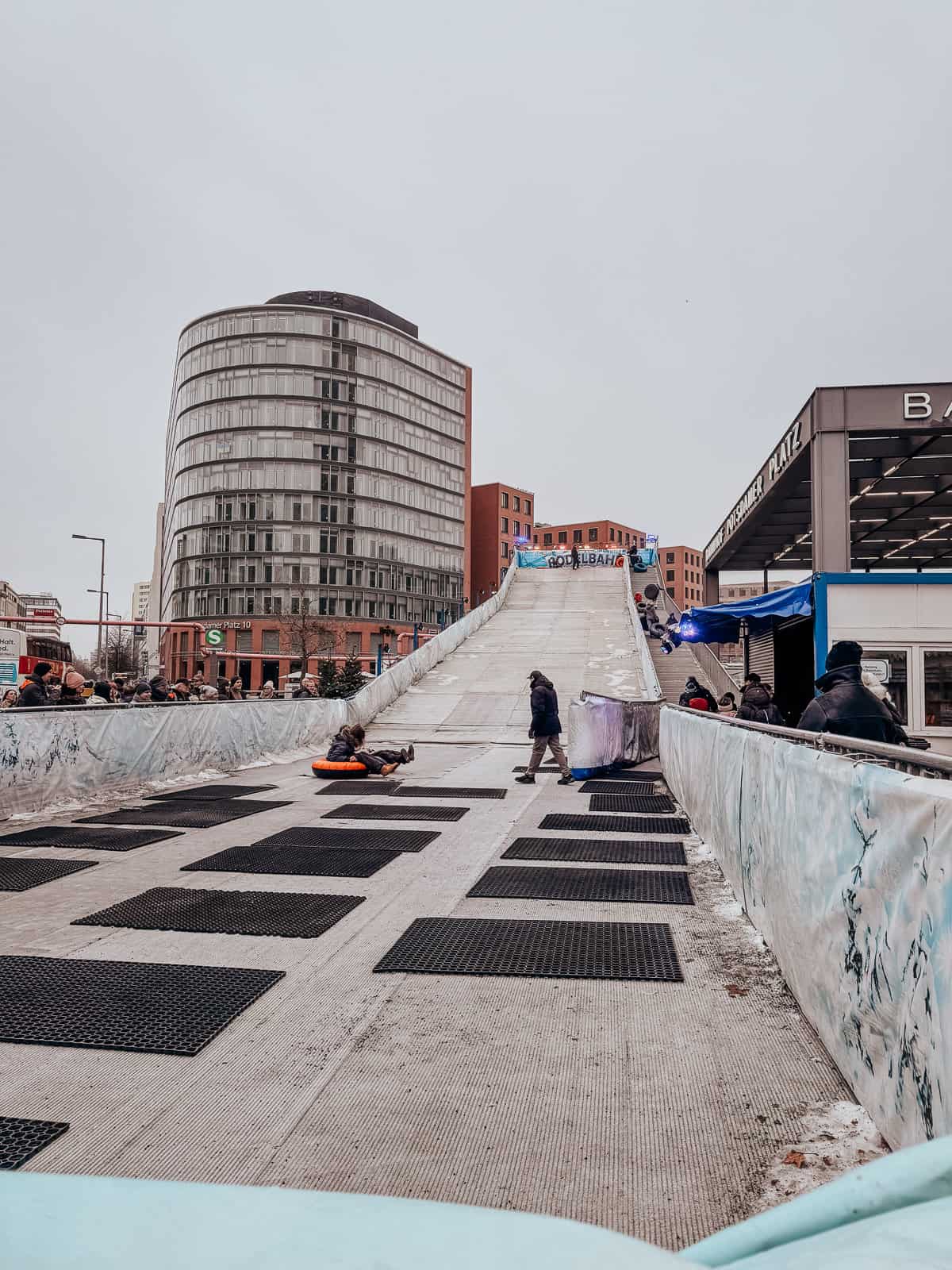 Children and adults enjoying a temporary urban snow slide, with inflatable rings. The slide is set amidst modern architecture, providing a unique blend of city life and winter fun
