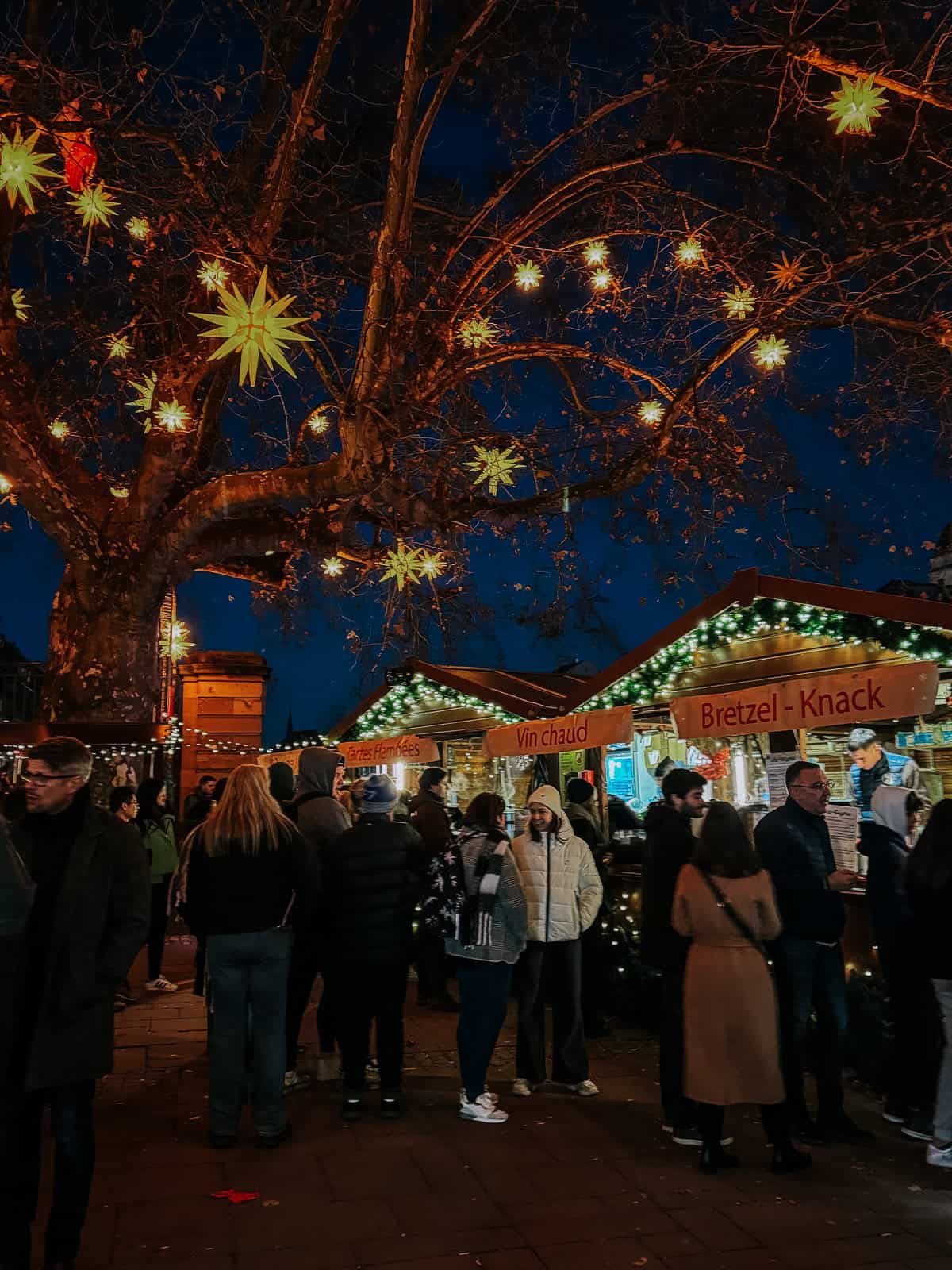 A vibrant Christmas market scene under a twilight sky, featuring an archway with the words 'Christkindelsmärik' illuminated by lights and surrounded by busy market sta