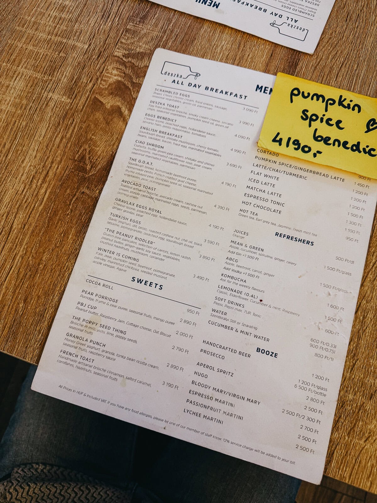 A cafe menu laid out on a wooden table, listing various breakfast items, beverages, and sweets, with a handwritten note highlighting "pumpkin spice benedict 4190 Ft.