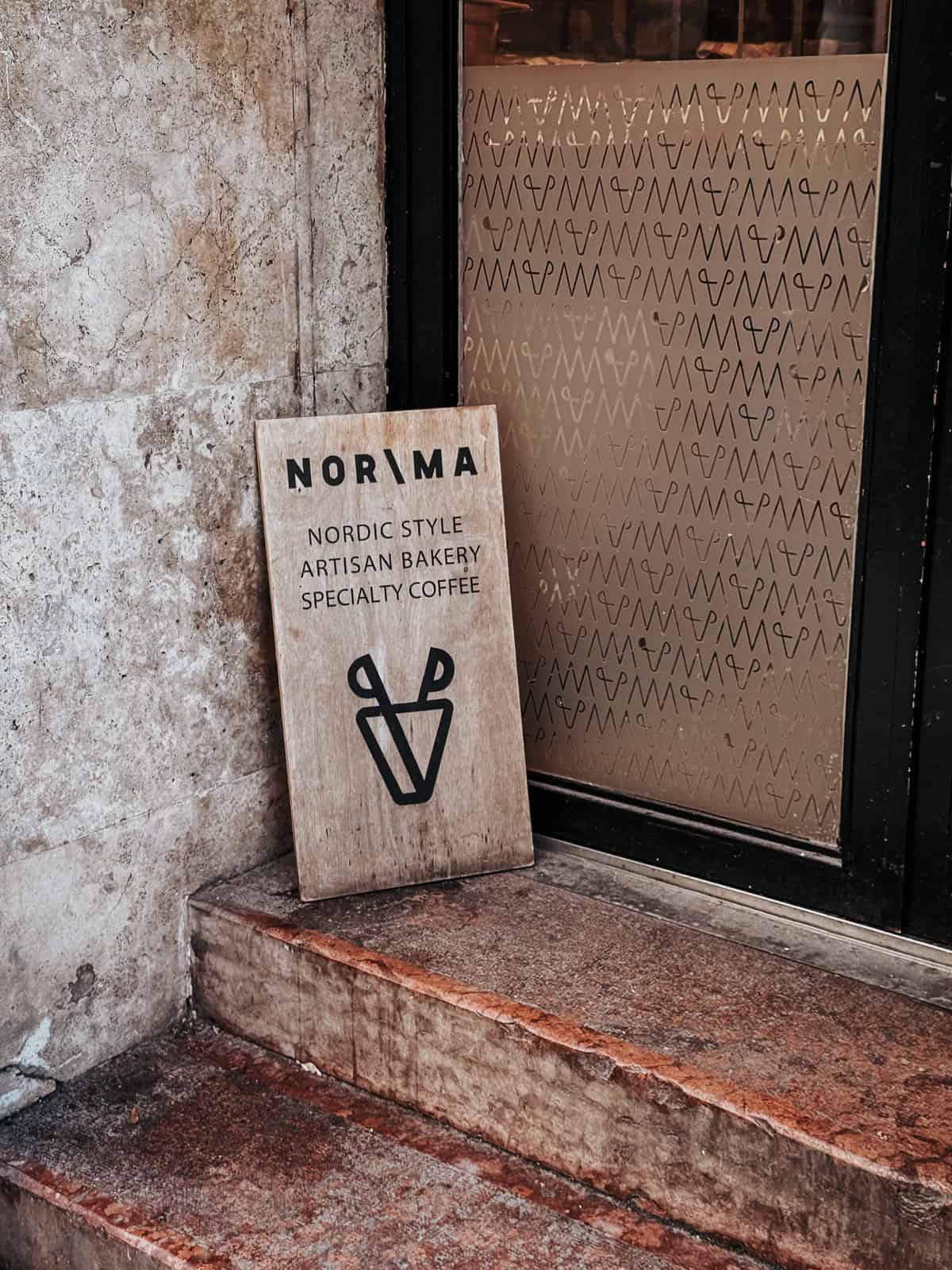 The entrance of Norma bakery with a signboard reading "NOR/MA Nordic Style Artisan Bakery Specialty Coffee" leaning against a textured wall, next to a glass door.