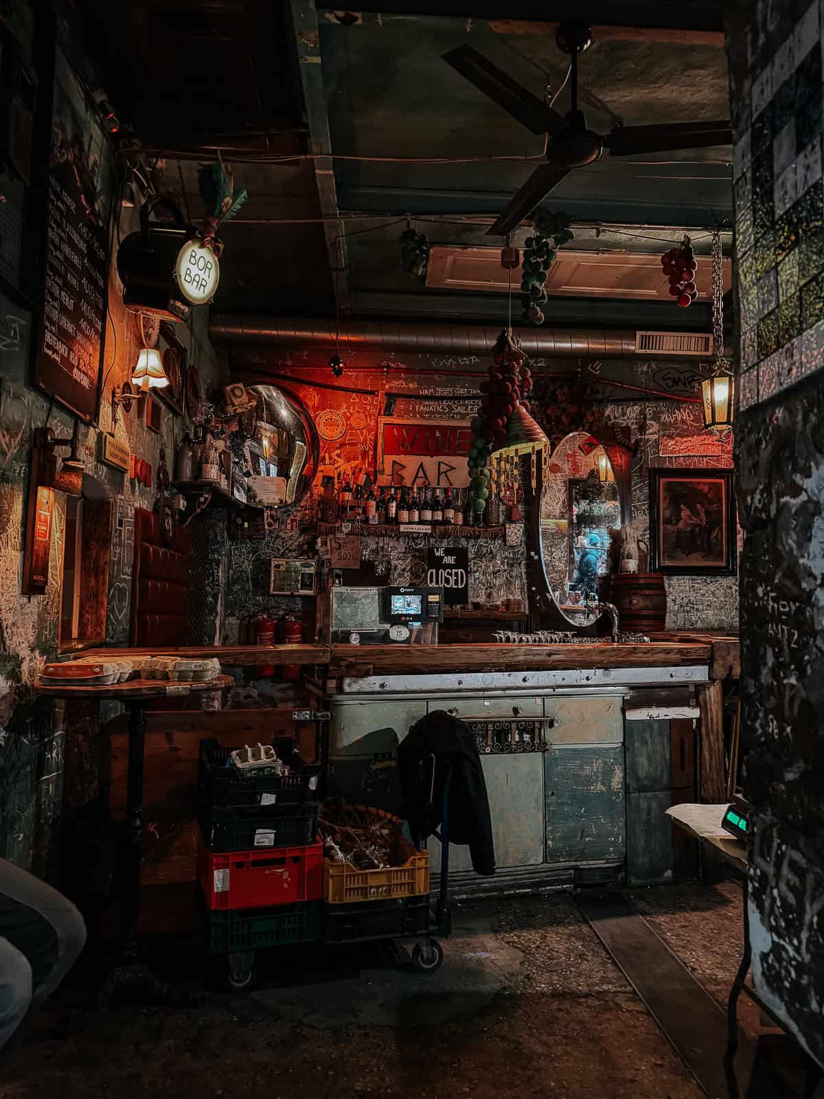 A dimly lit, rustic bar with a cozy atmosphere, featuring a vintage bar counter, hanging grapes decor, and a sign that reads "Wine Bar" on the back wall
