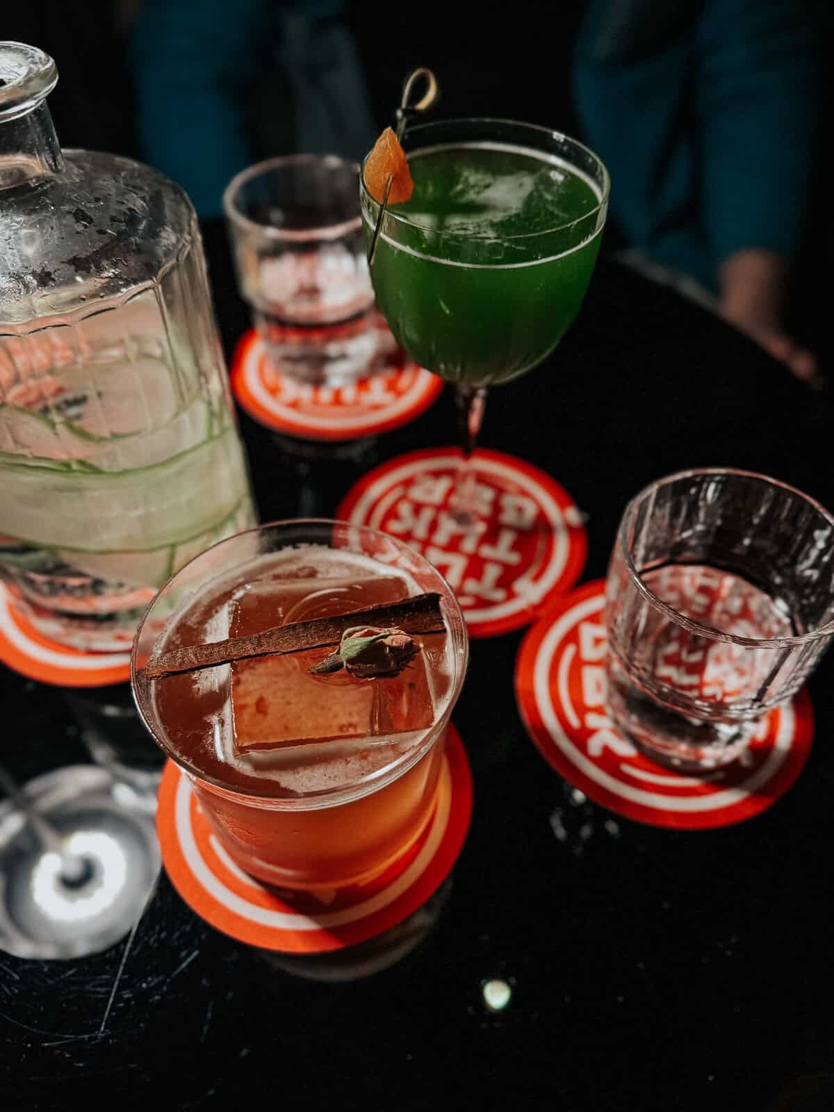 A close-up shot of colorful cocktails on a black table, with glasses and a water pitcher, all placed on vibrant red coasters.