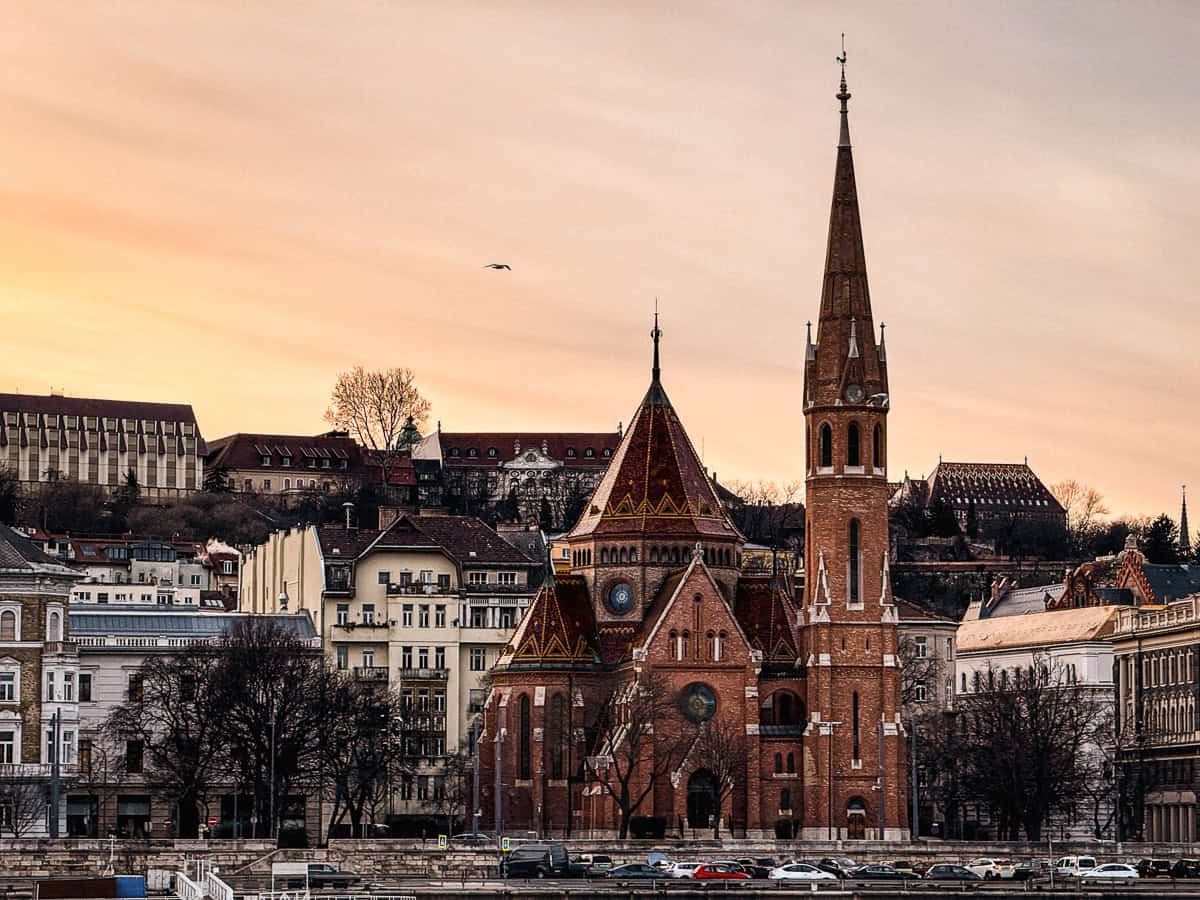 A scenic view of a historic church with intricate architecture, surrounded by other old buildings, under a soft orange sunset sky.