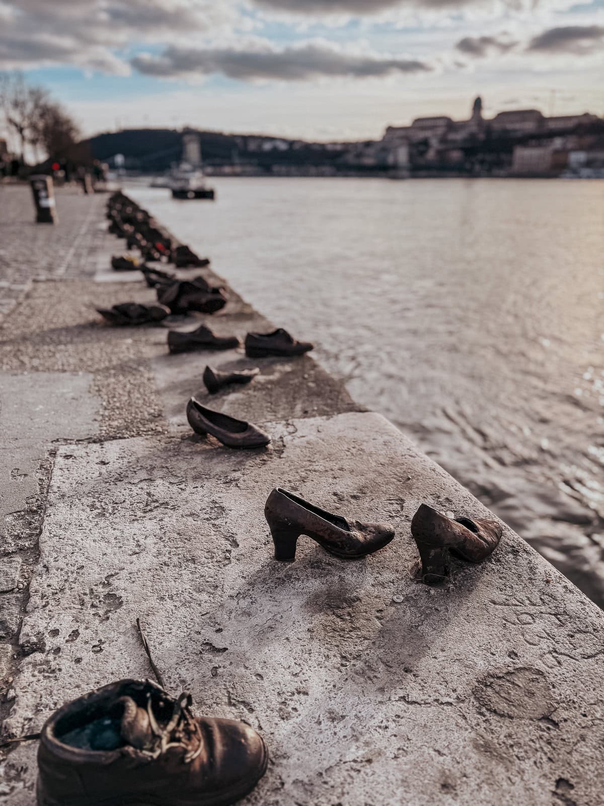 The "Shoes on the Danube Bank" memorial in Budapest, with scattered old shoes along the riverside commemorating the Holocaust victims, with the river and city in the background.