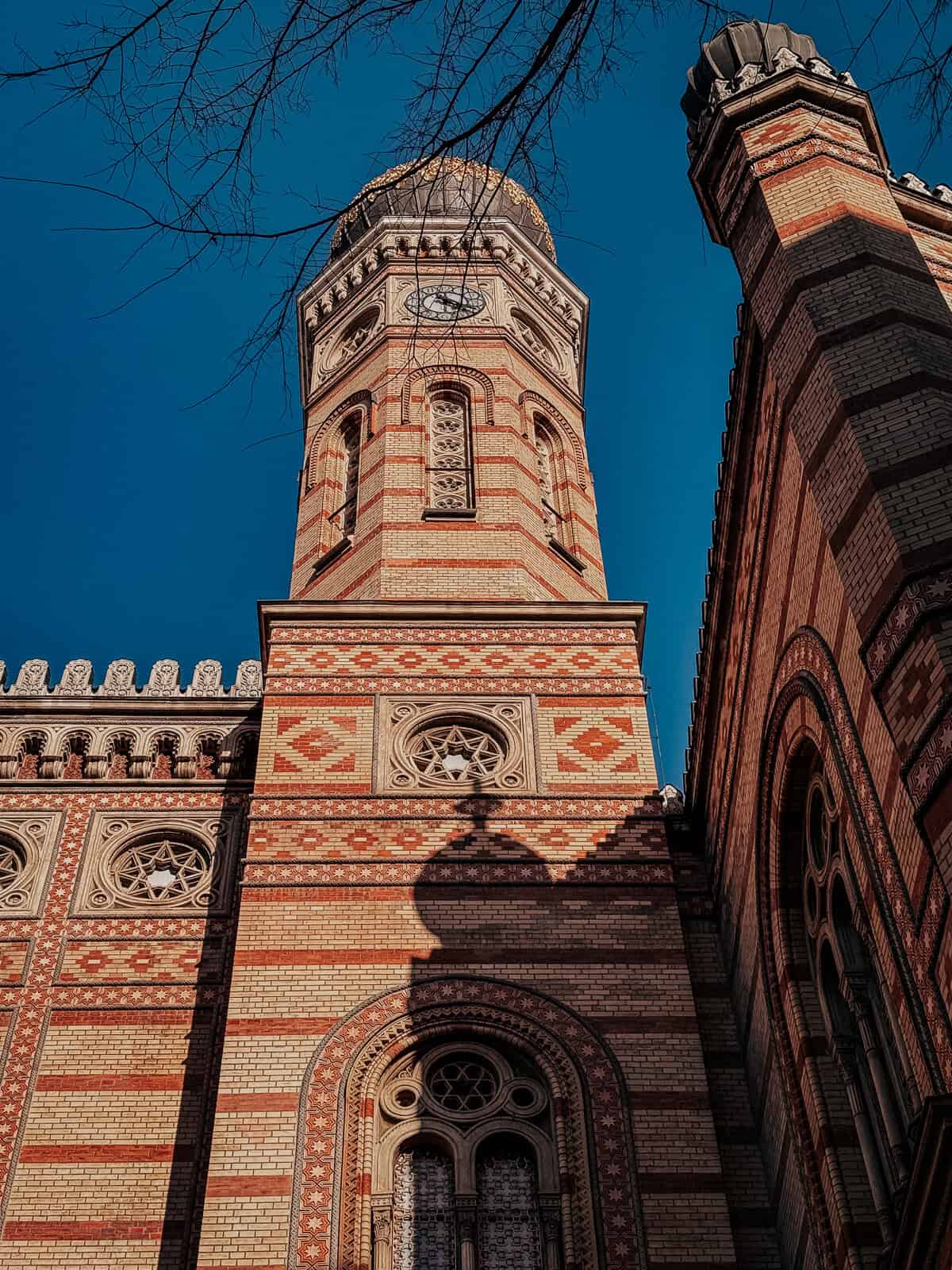A low-angle shot of a tall clock tower of a grand building, showing detailed brickwork and architectural features, under a clear blue sky.