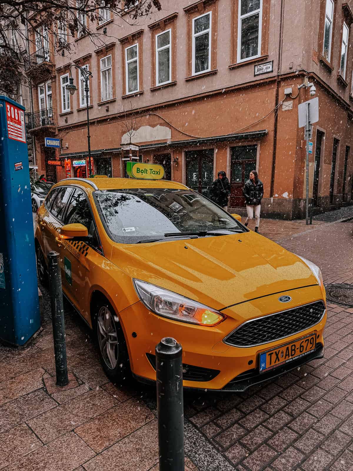 A yellow Bolt taxi parked on a snowy street in Budapest, with a "Change" currency exchange sign and pedestrians in the background.