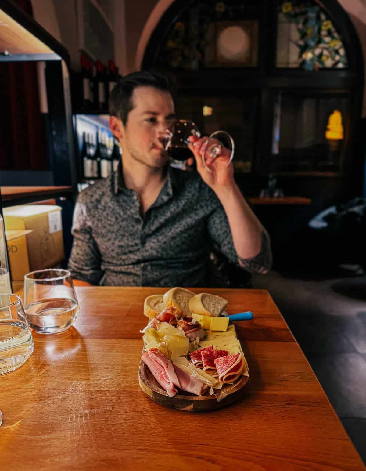 A man in a patterned shirt is sitting at a wooden table, sipping red wine. A charcuterie board with various meats, cheeses, and bread is in front of him.