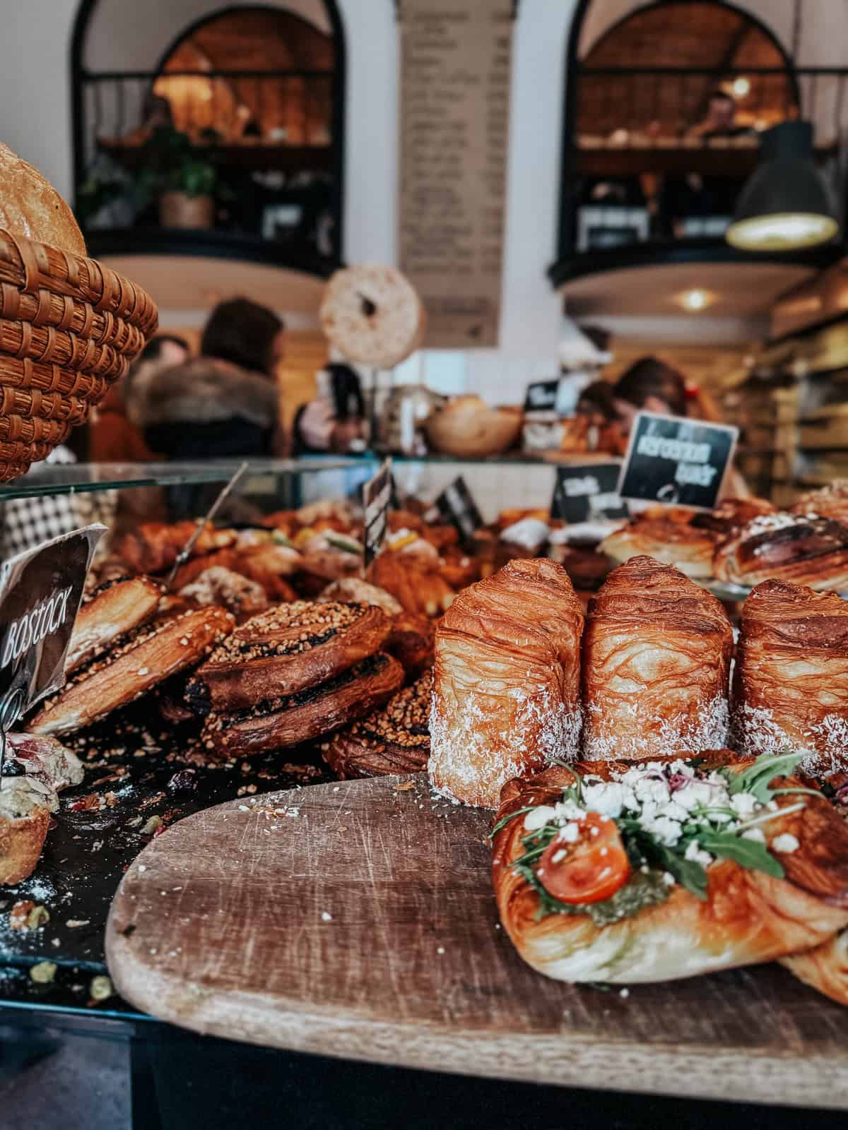 An assortment of pastries and baked goods is displayed on a wooden board inside a bakery, with various pastries labeled and customers in the background.