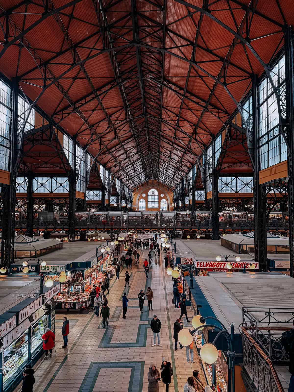 An interior view of Budapest Central Market Hall, showcasing its high, arched ceilings and rows of market stalls with people browsing and shopping.