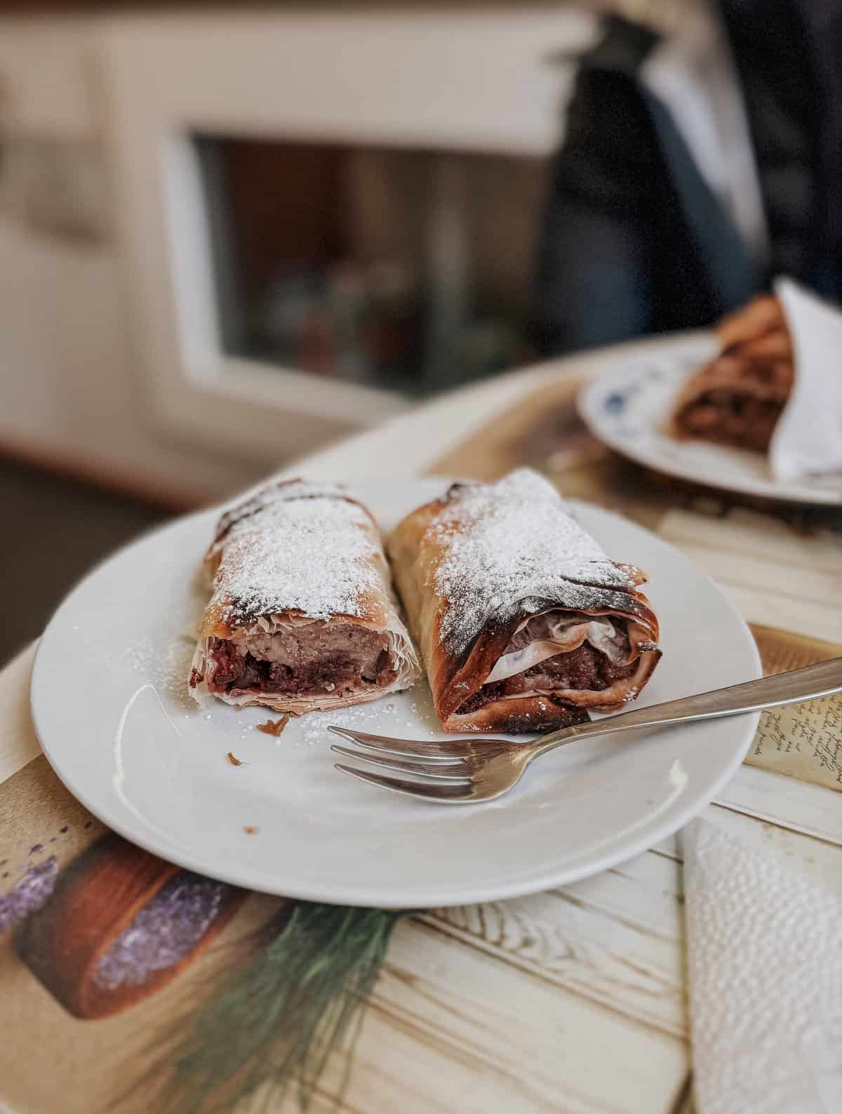 A close-up of two slices of strudel on a white plate, dusted with powdered sugar and accompanied by a fork.