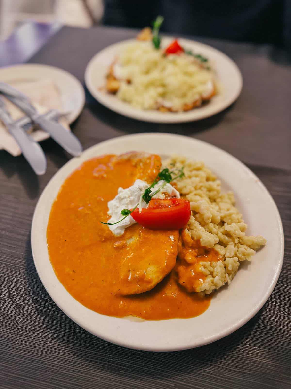 A plate of chicken paprikash with creamy paprika sauce, garnished with a tomato slice and herbs, served with a side of dumplings.