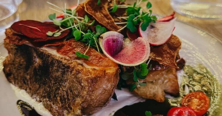 A plate of savory dish with thick bread, garnished with vibrant greens, cherry tomatoes, and radish slices, on a wooden table with a person in the background.