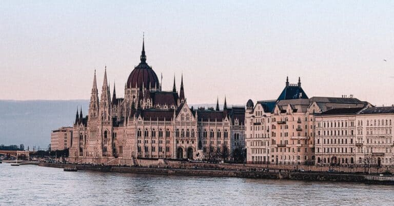 A view of the Hungarian Parliament Building from across the river, highlighting its gothic architecture and domes under a clear sky.