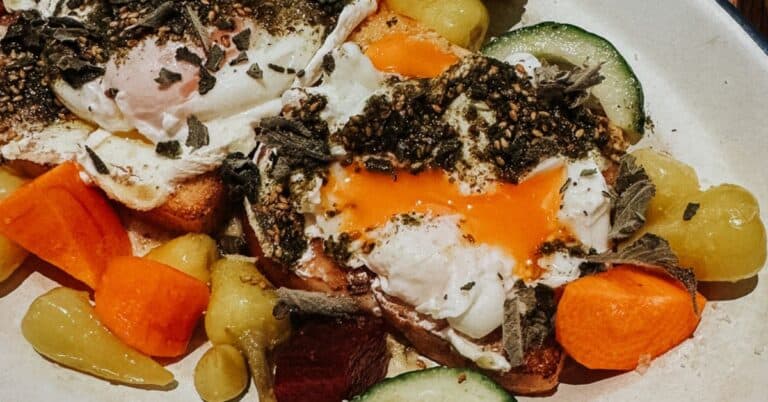 A colorful brunch plate with two eggs, diced vegetables, and a mix of pickles, set on a wooden table with a glass of juice.