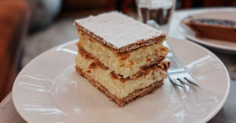 A slice of Hungarian kremes, a layered pastry with cream filling, served on a white plate in a cafe, with a glass of water and a blurred background of a person sitting.