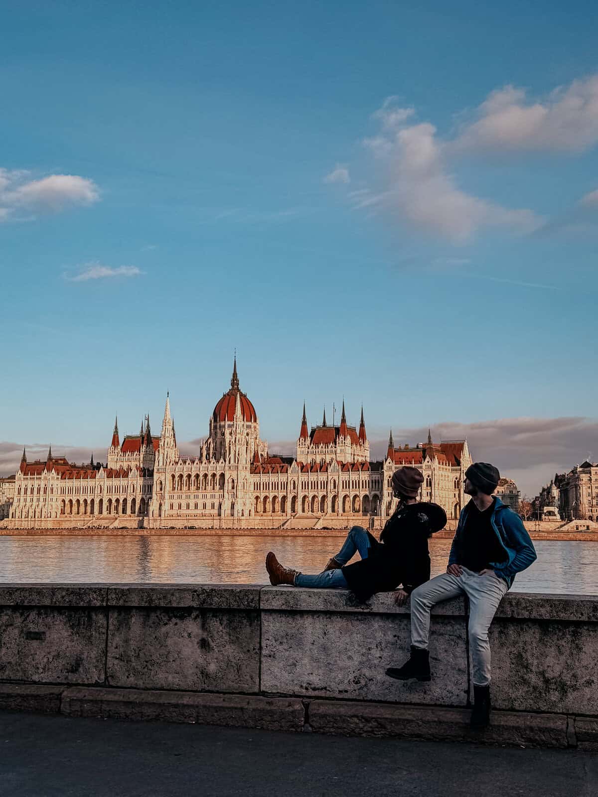 Two people sit on a stone ledge by the river, with the Hungarian Parliament Building in the background during sunset, reflecting warm light off the building.