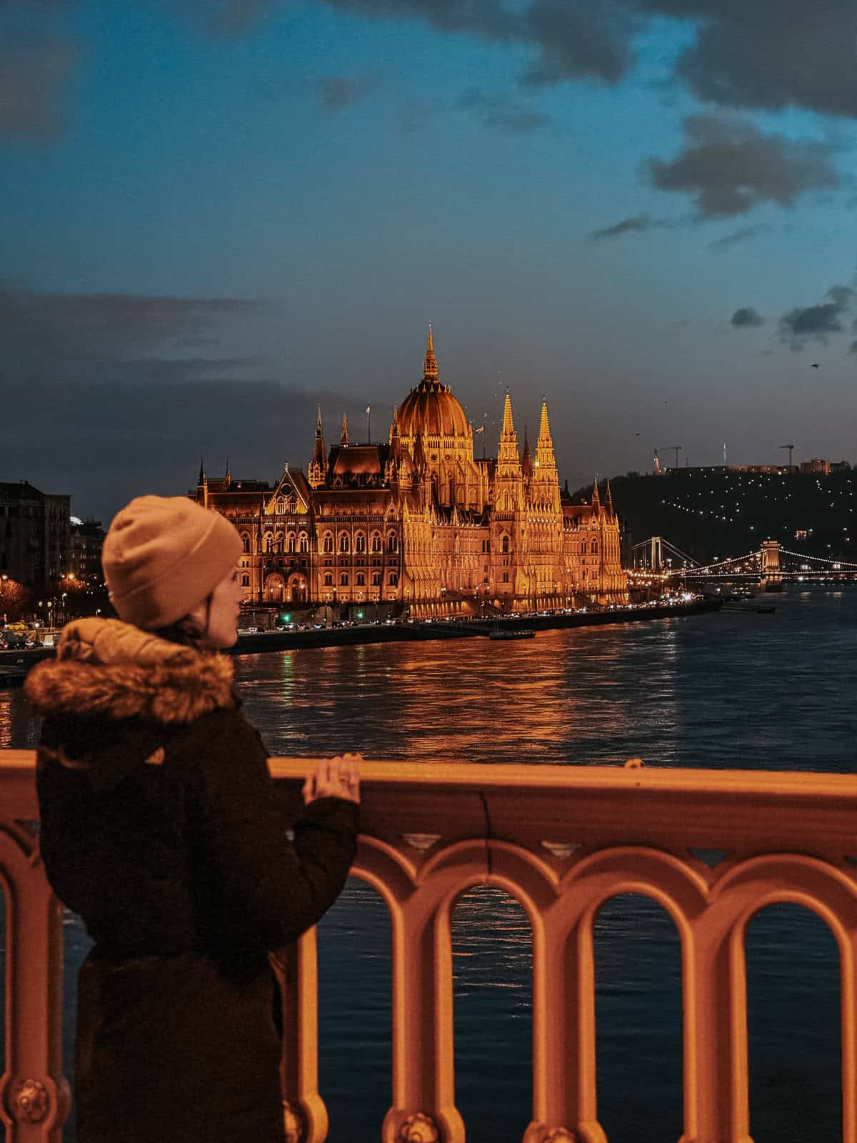 A woman in a pink beanie and black coat stands on a bridge, gazing at the illuminated Hungarian Parliament Building at night, reflecting on the river.