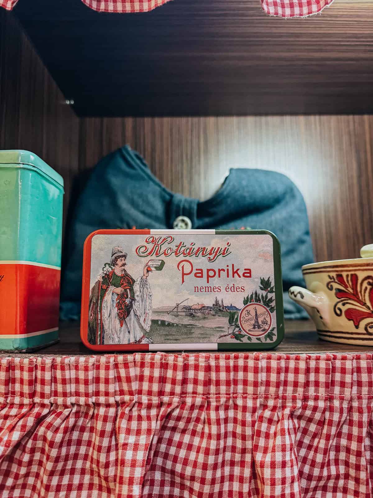 A vintage-style tin of Hungarian paprika, labeled "Kotányi Paprika," displayed on a shelf with red-and-white checkered fabric.