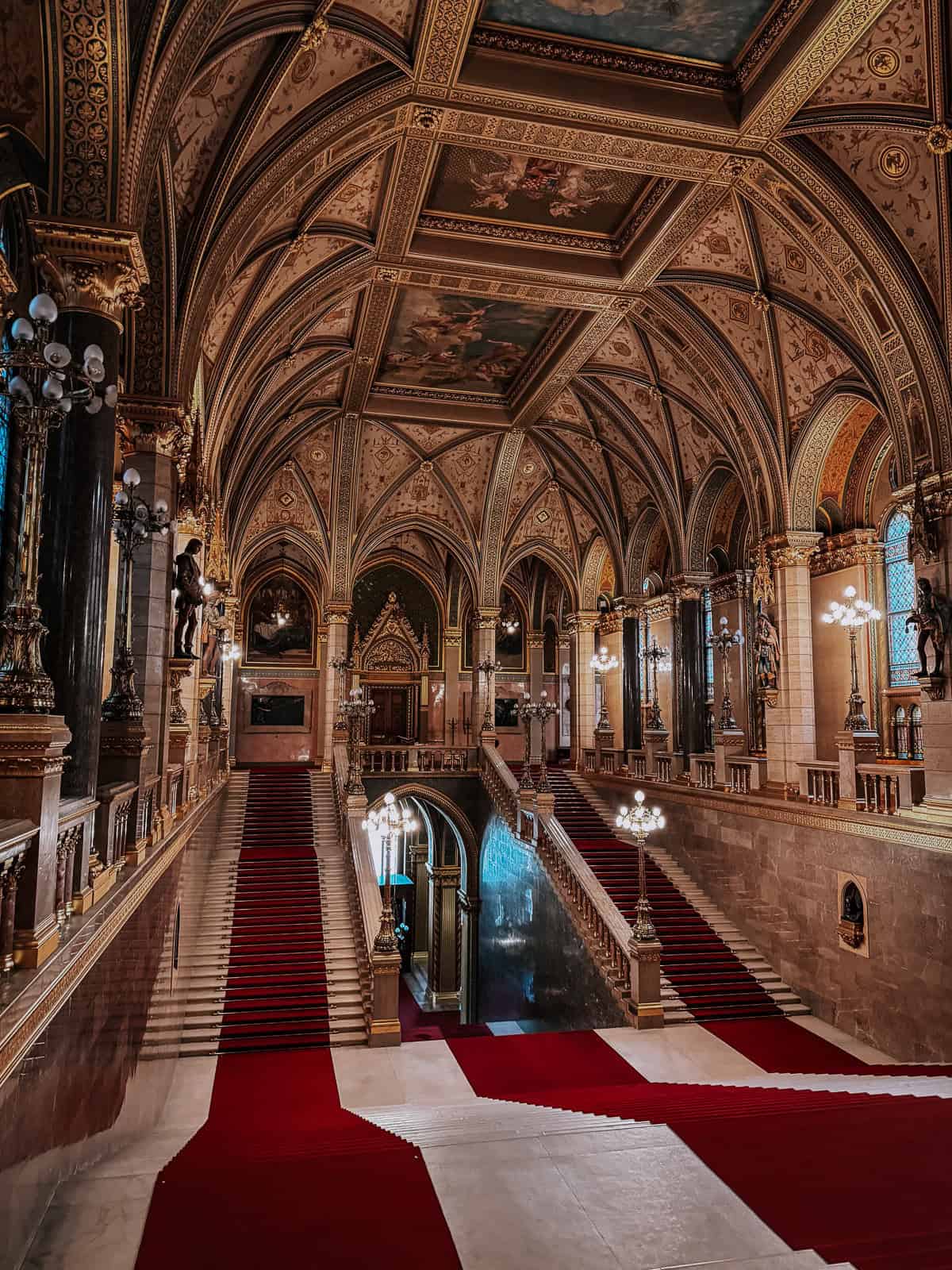 The grand interior of the Hungarian Parliament, featuring ornate ceilings, red carpeted stairs, and elaborate decorations with intricate lighting fixtures.