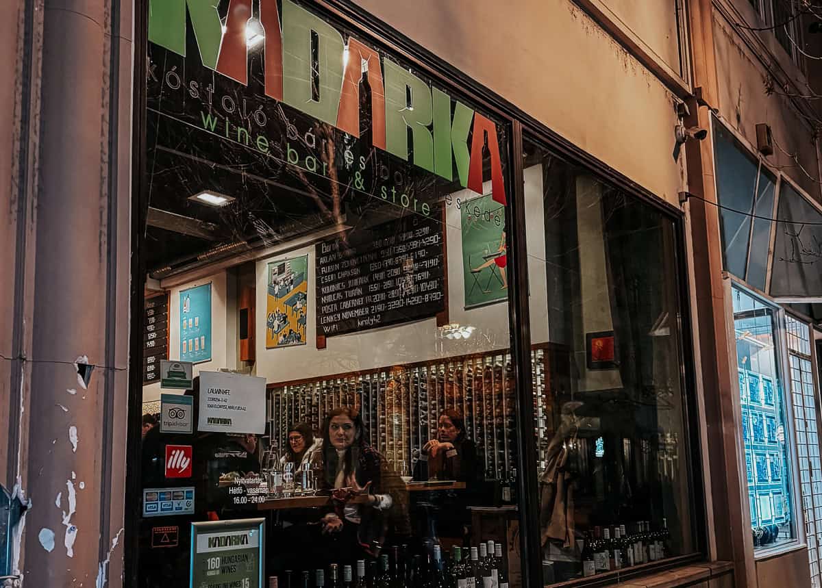 The exterior of a wine bar named "Kadarka," with large windows showing people inside enjoying their drinks. The name is prominently displayed in green and red letters above the window.