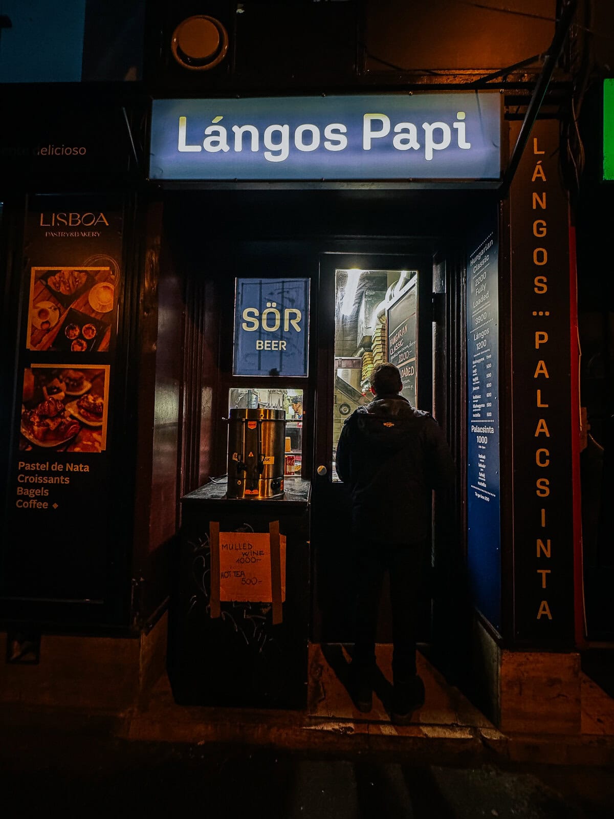 The entrance to a small restaurant named "Lángos Papi," with a brightly lit sign. The restaurant offers beer, and a small handwritten sign on a board mentions mulled wine and hot tea prices.