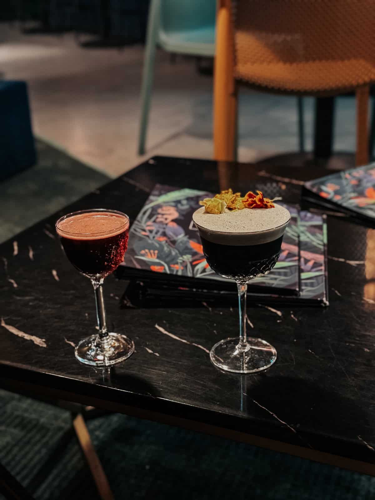 Two beautifully garnished cocktails on a dark marble table, set against a backdrop of modern, colorful decor."