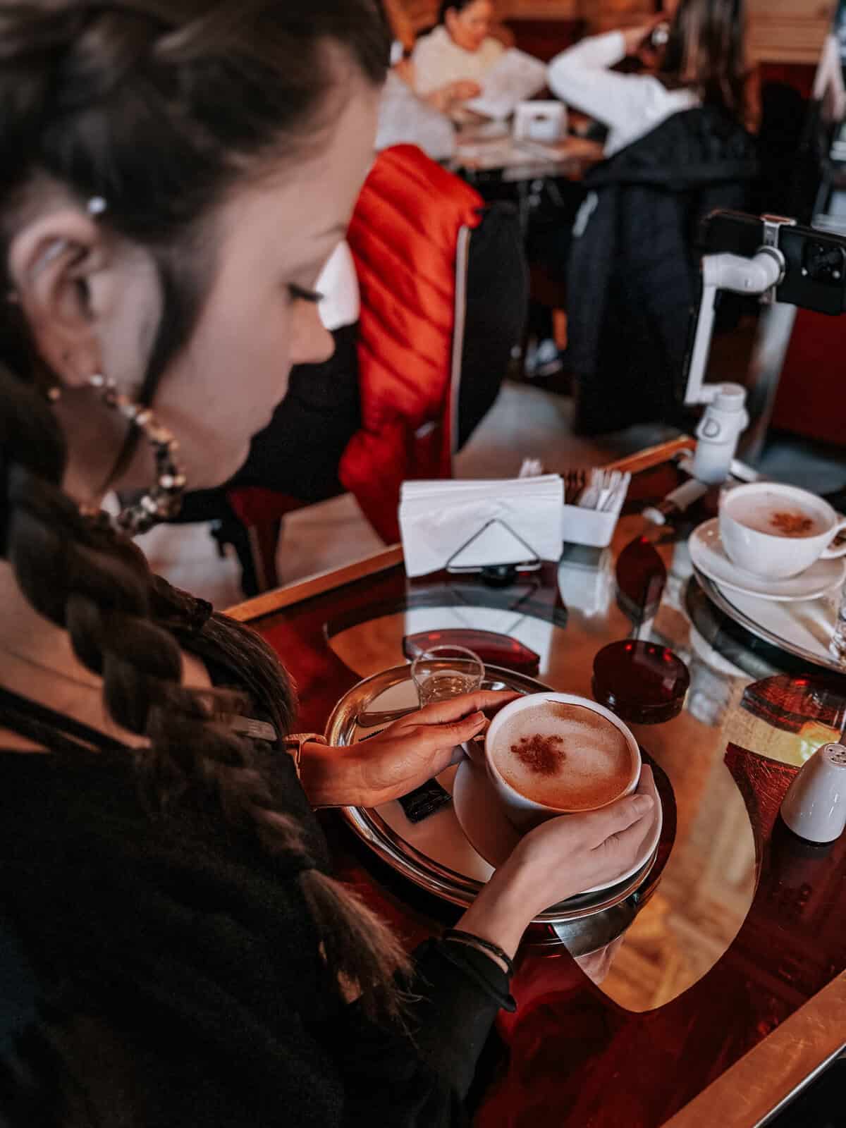 A woman with braided hair and large hoop earrings enjoys a coffee at the New York Cafe, holding a cup of frothy coffee at a table with a red reflective surface.