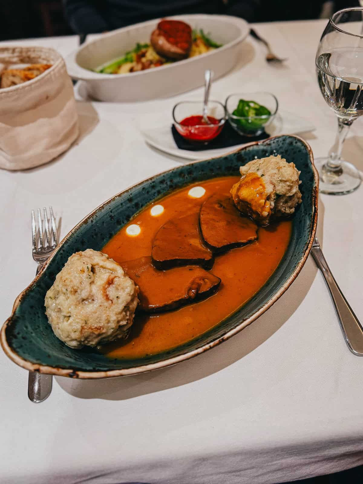 Plate of traditional Hungarian beef stew with bread dumplings, served in a rich, flavorful sauce, accompanied by condiments and a glass of white wine on a white tablecloth.