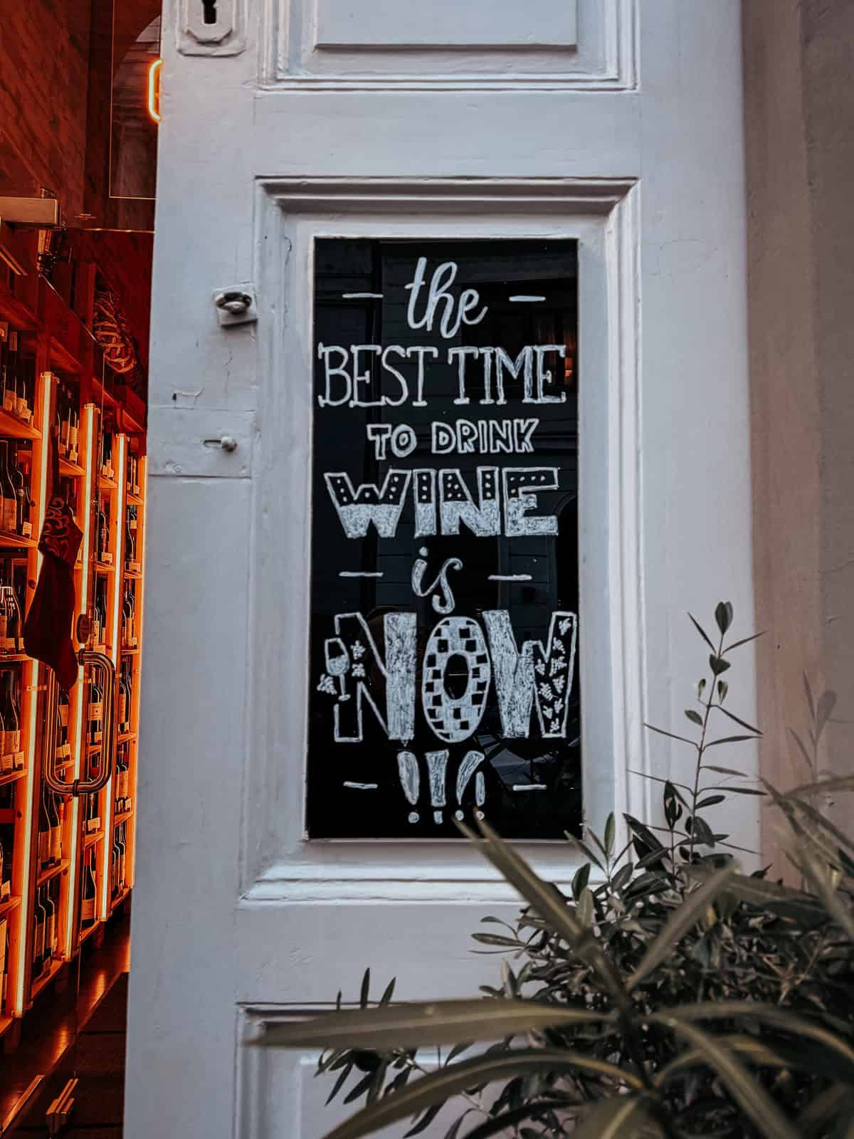 sign on a door reads "The best time to drink wine is now!!!" with a cozy wine shop interior visible through the glass.