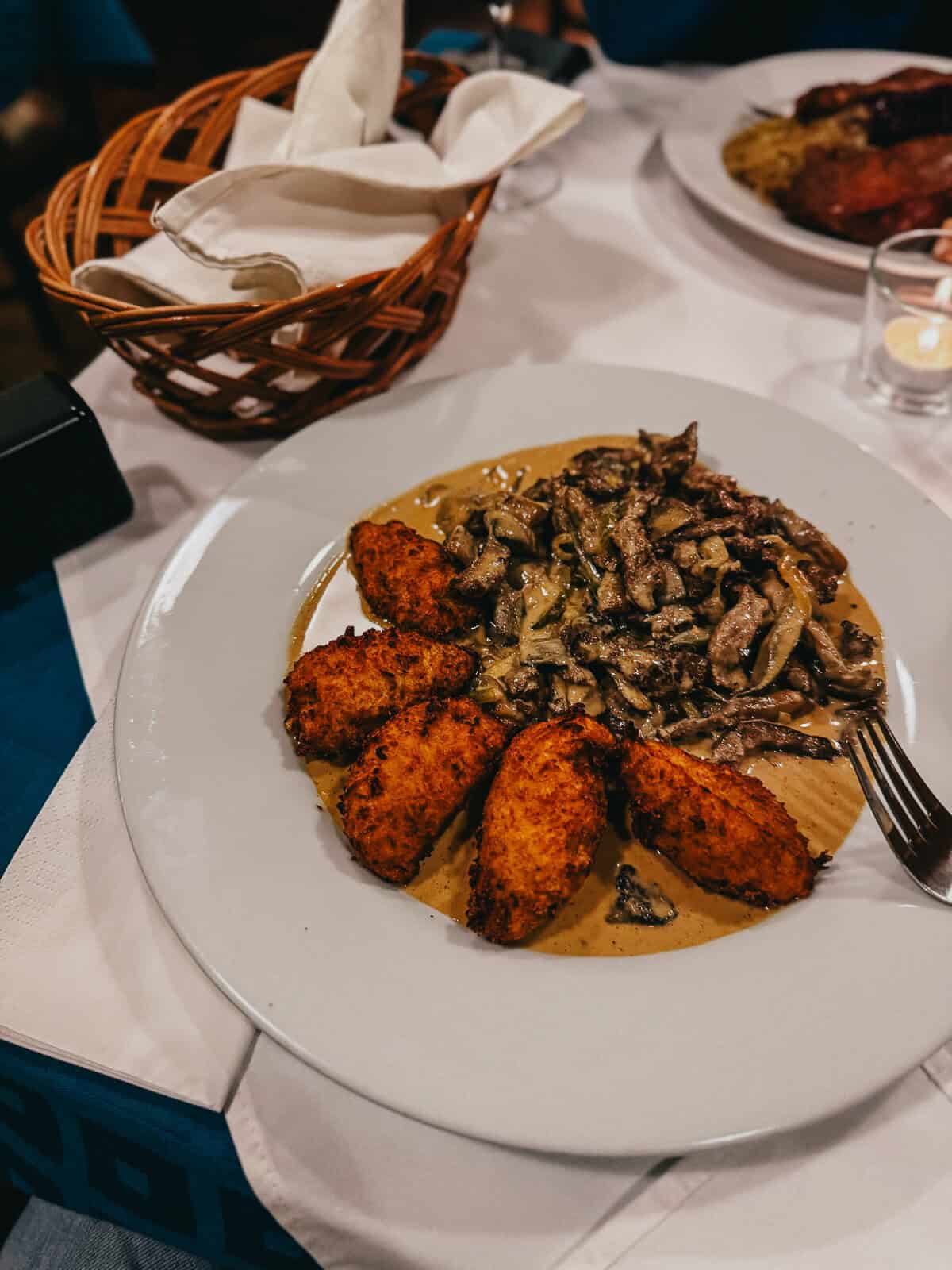 A plate of fried croquettes and a creamy mushroom sauce served at a restaurant, with a basket of bread and another dish in the background.
