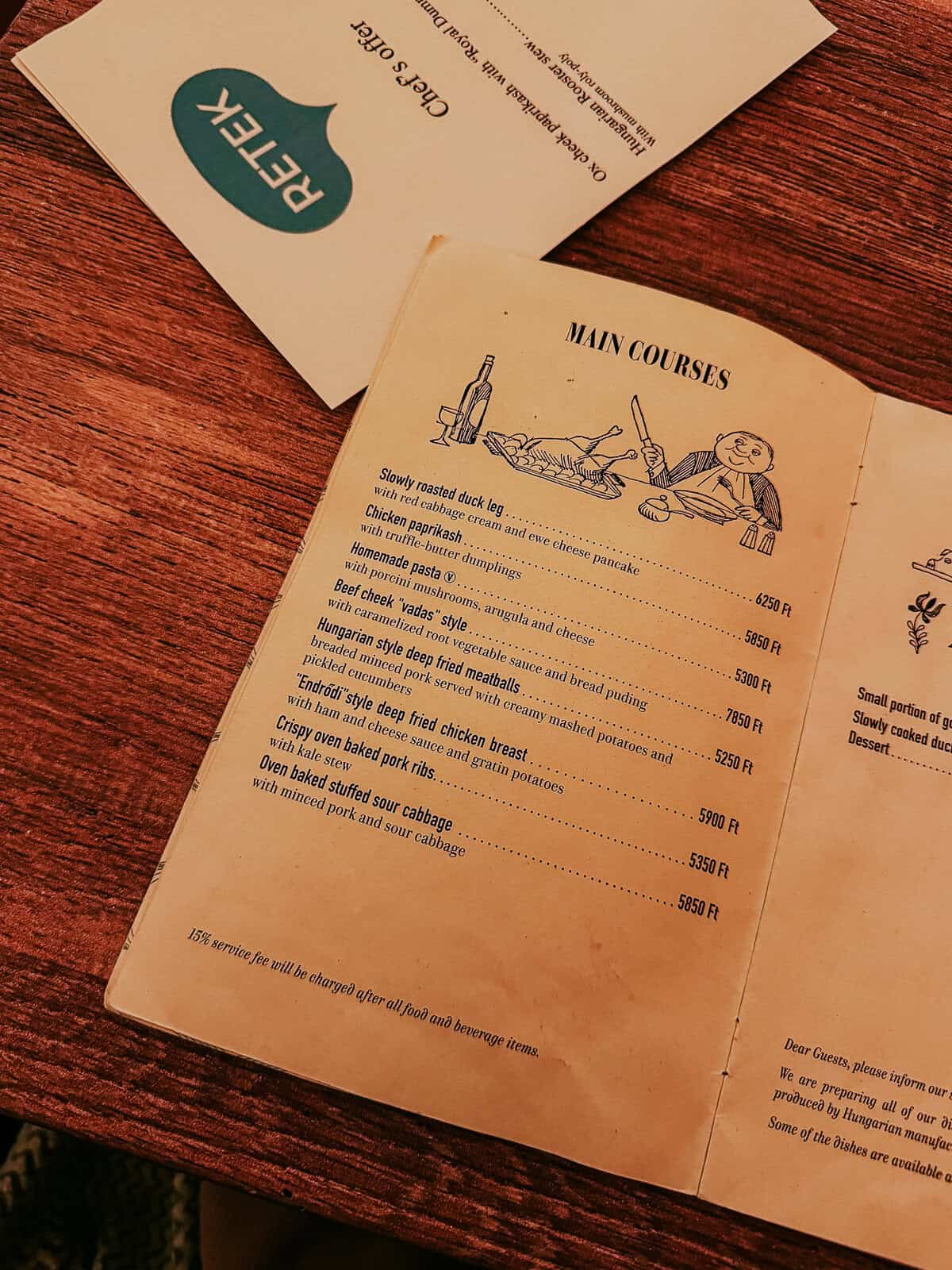 An open menu on a wooden table listing various main courses, including "Slowly roasted duck leg" and "Chicken paprikash with truffle-butter dumplings."