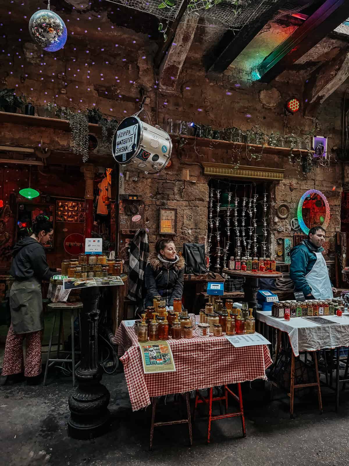 A cozy ruin bar setting with eclectic decorations, including jars of honey on tables covered with checkered tablecloths, and colorful lights hanging from the ceiling.
