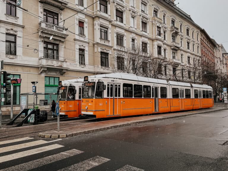 Two orange trams on the street in Budapest, with a historic building in the background and people waiting at the tram stop.