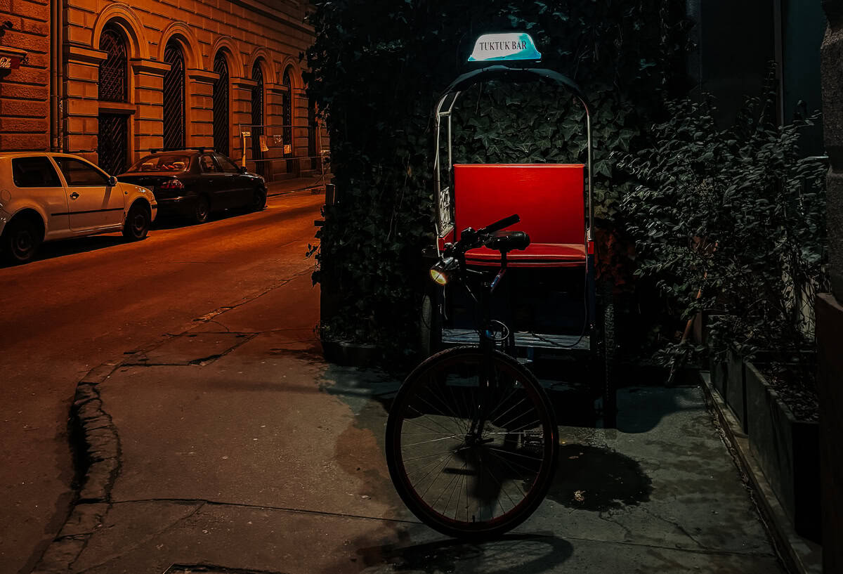 A rickshaw with a sign reading "Tuk Tuk Bar" parked on a quiet street at night. The rickshaw has a red seat and is partially covered with ivy, creating a cozy and secluded feel.