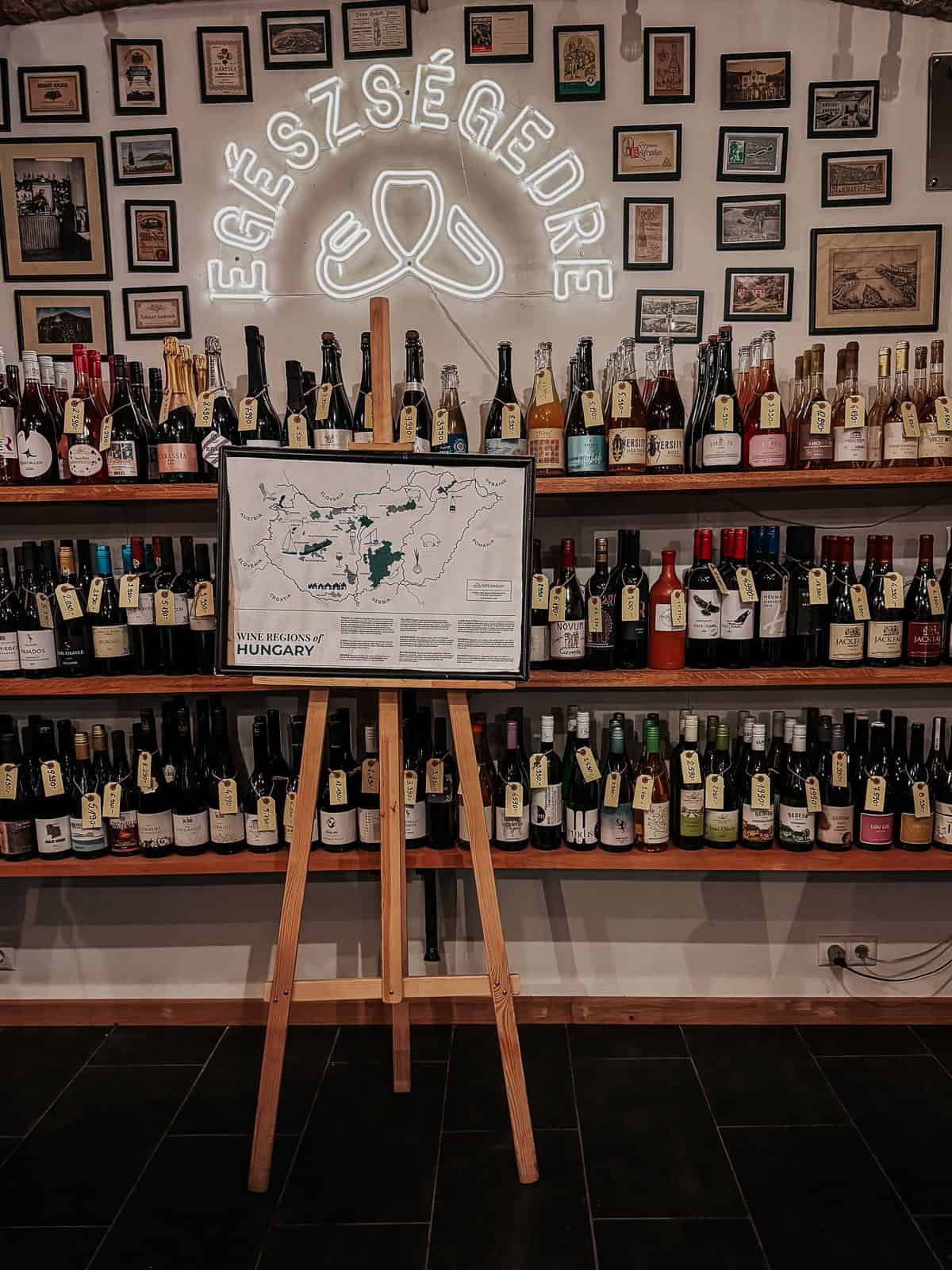A wine shop interior with shelves full of various wine bottles. A neon sign with the word "Egészségedre" (meaning "Cheers" in Hungarian) and a map of wine regions in Hungary are prominently displayed.