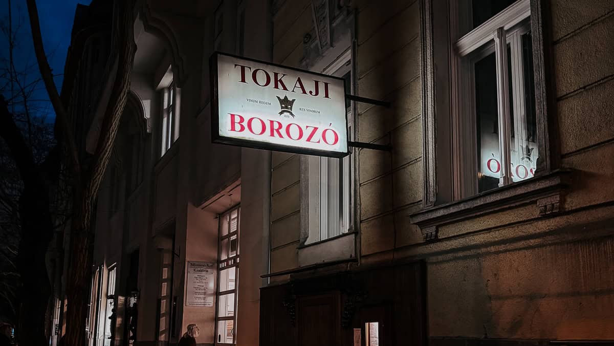 An exterior night view of a building with a lit sign reading "Tokaji Borozó." The sign and surrounding architecture are warmly illuminated against the dark evening backdrop.