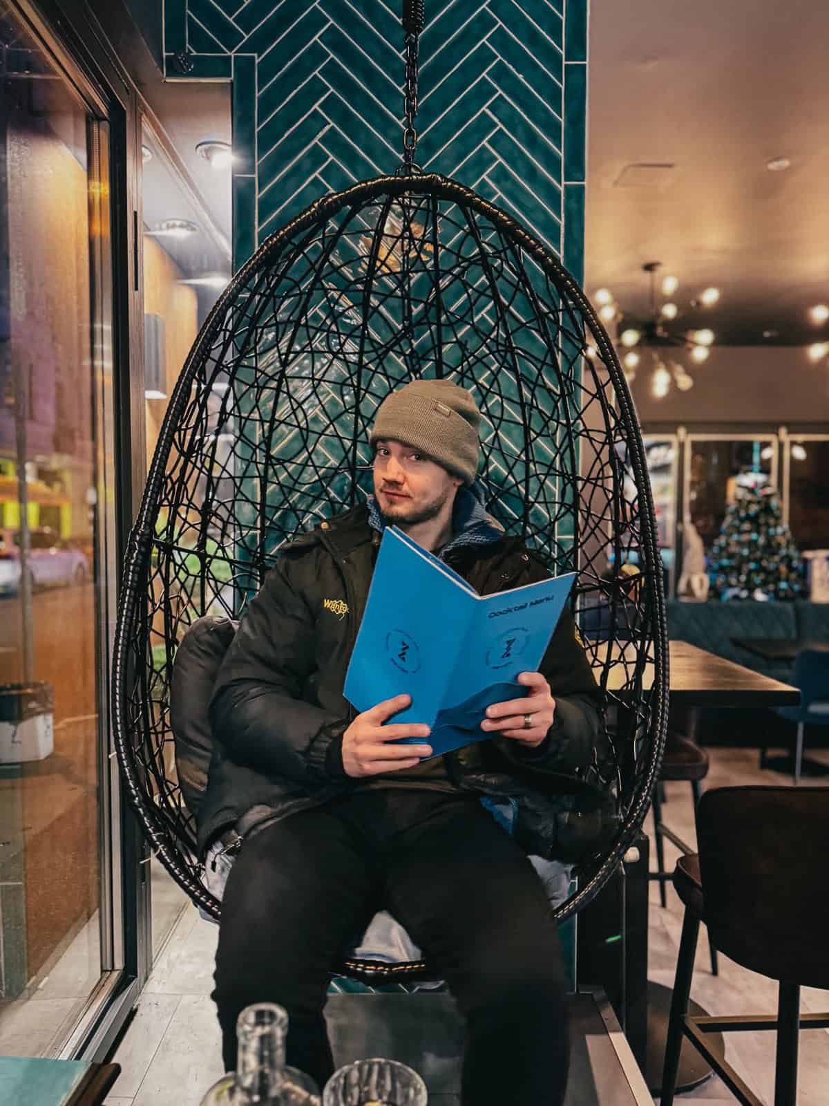A person sitting in a hanging wicker chair inside a bar, reading a blue menu, with a green tiled wall behind."