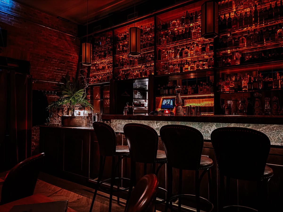 A dimly lit bar with dark wooden furniture and shelves filled with various spirits, creating a warm and intimate atmosphere."