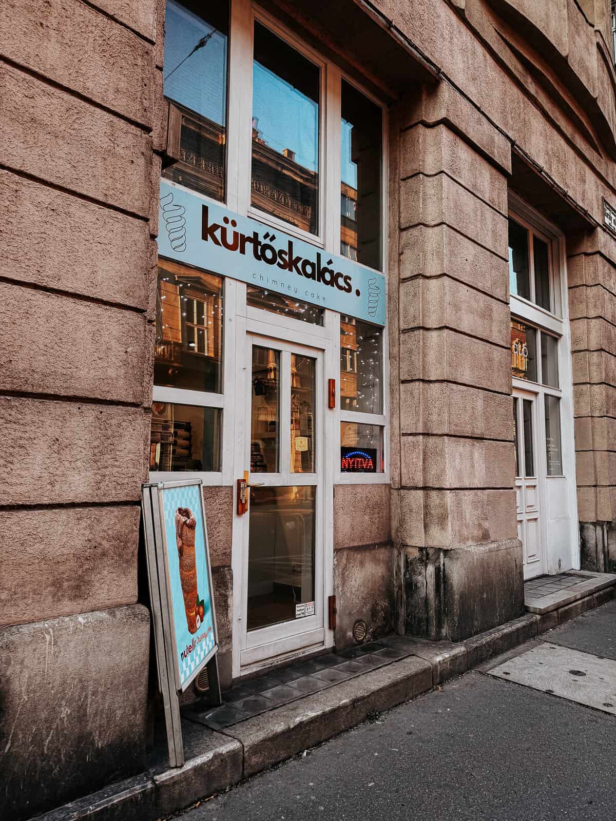 The exterior of a shop with a sign that reads "Kürtőskalács" (chimney cake) above the door. A poster of a chimney cake is displayed on a stand next to the entrance, which is located in a building with stone walls.