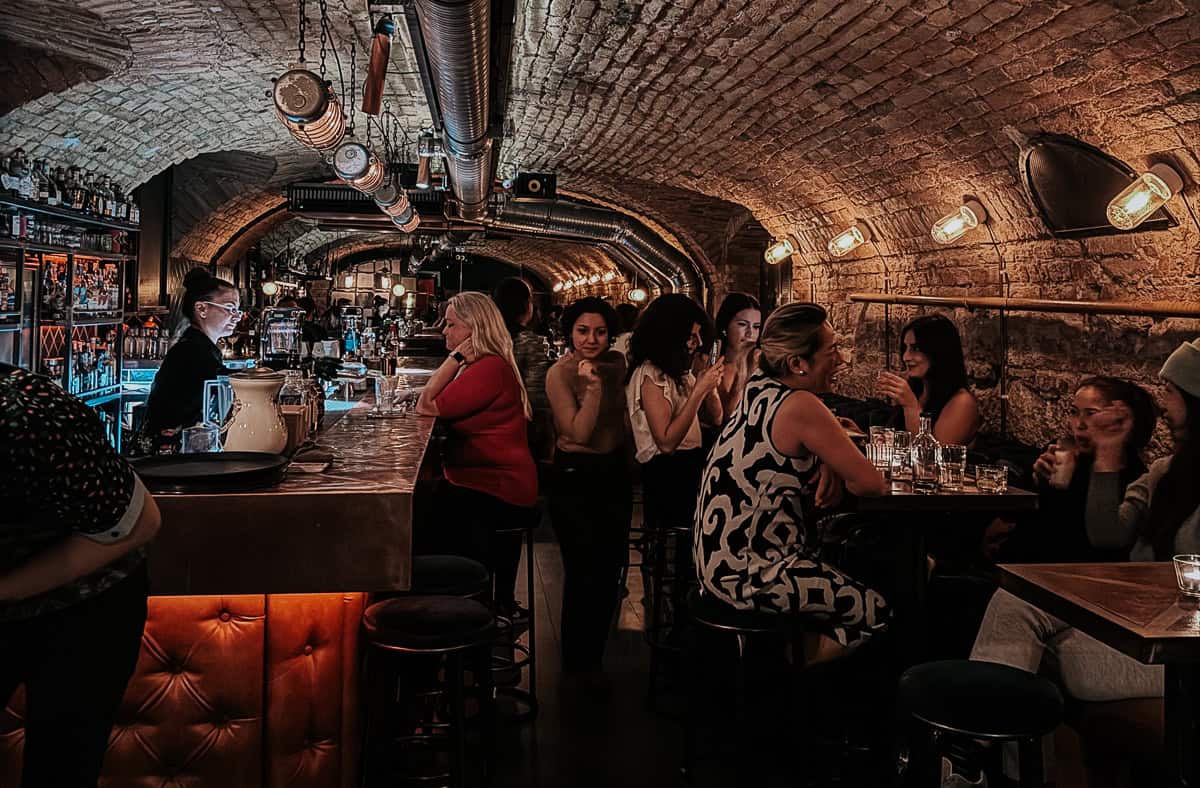 A cozy bar scene with patrons seated at tables and along the bar, featuring an arched brick ceiling and warm lighting."