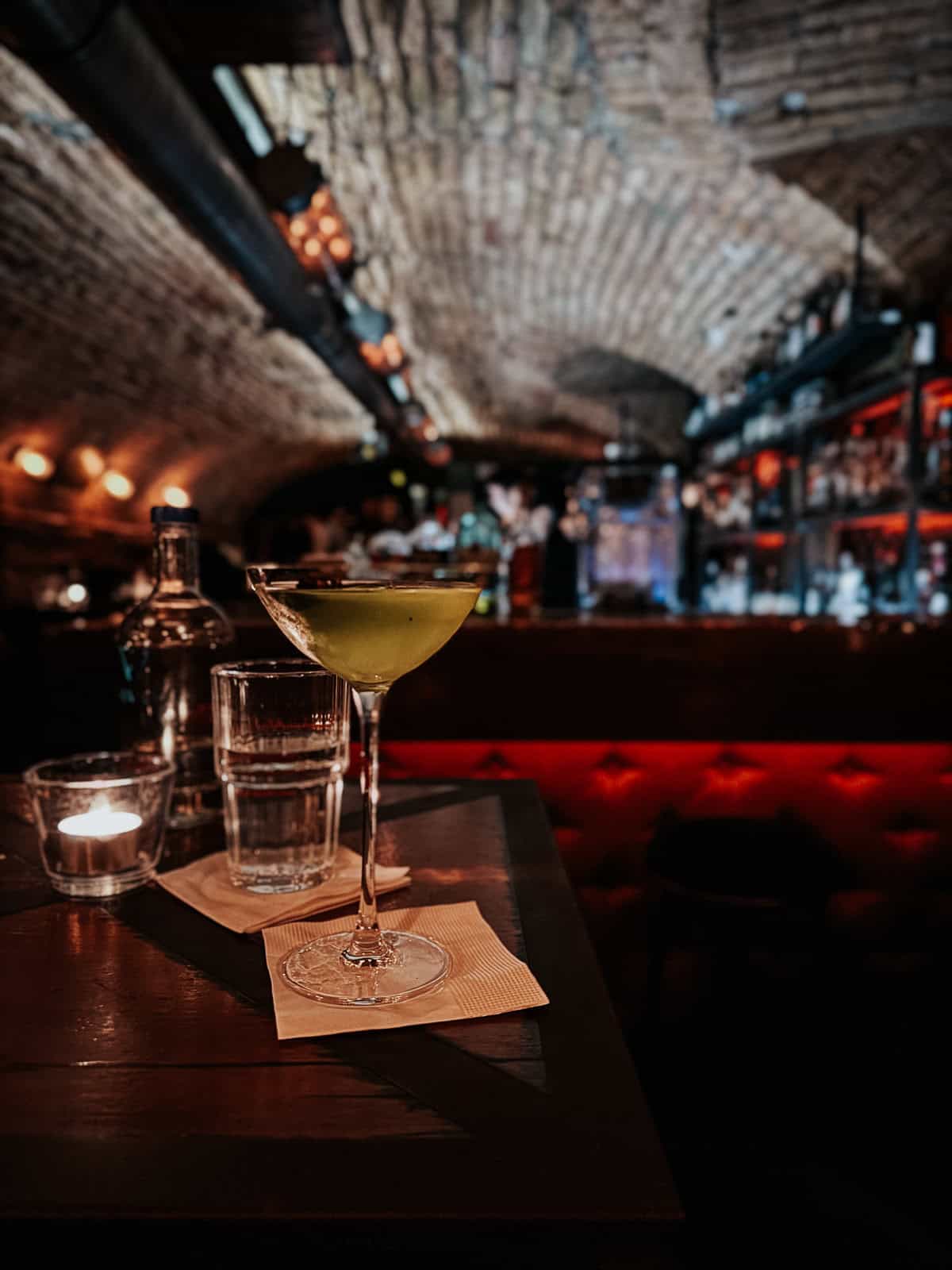 A cocktail served in a martini glass on a small table, with a candle and water glasses, set against a backdrop of an arched brick ceiling and softly lit bar."