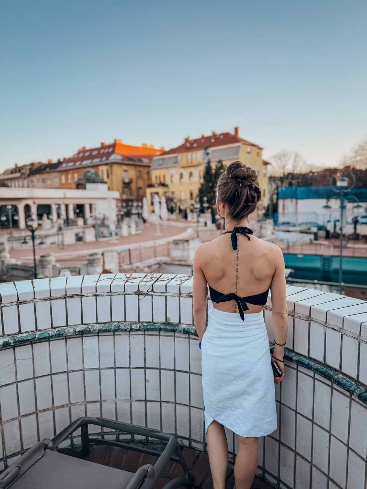 A woman in a black bikini top and white towel stands on a tiled balcony, overlooking the Széchenyi Thermal Bath in Budapest with historic buildings in the background.