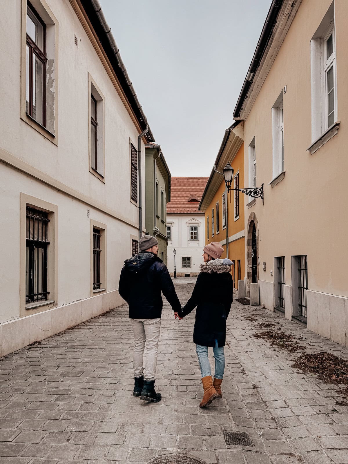 A couple holding hands, walking down a narrow cobblestone street lined with colorful historic buildings in Budapest.