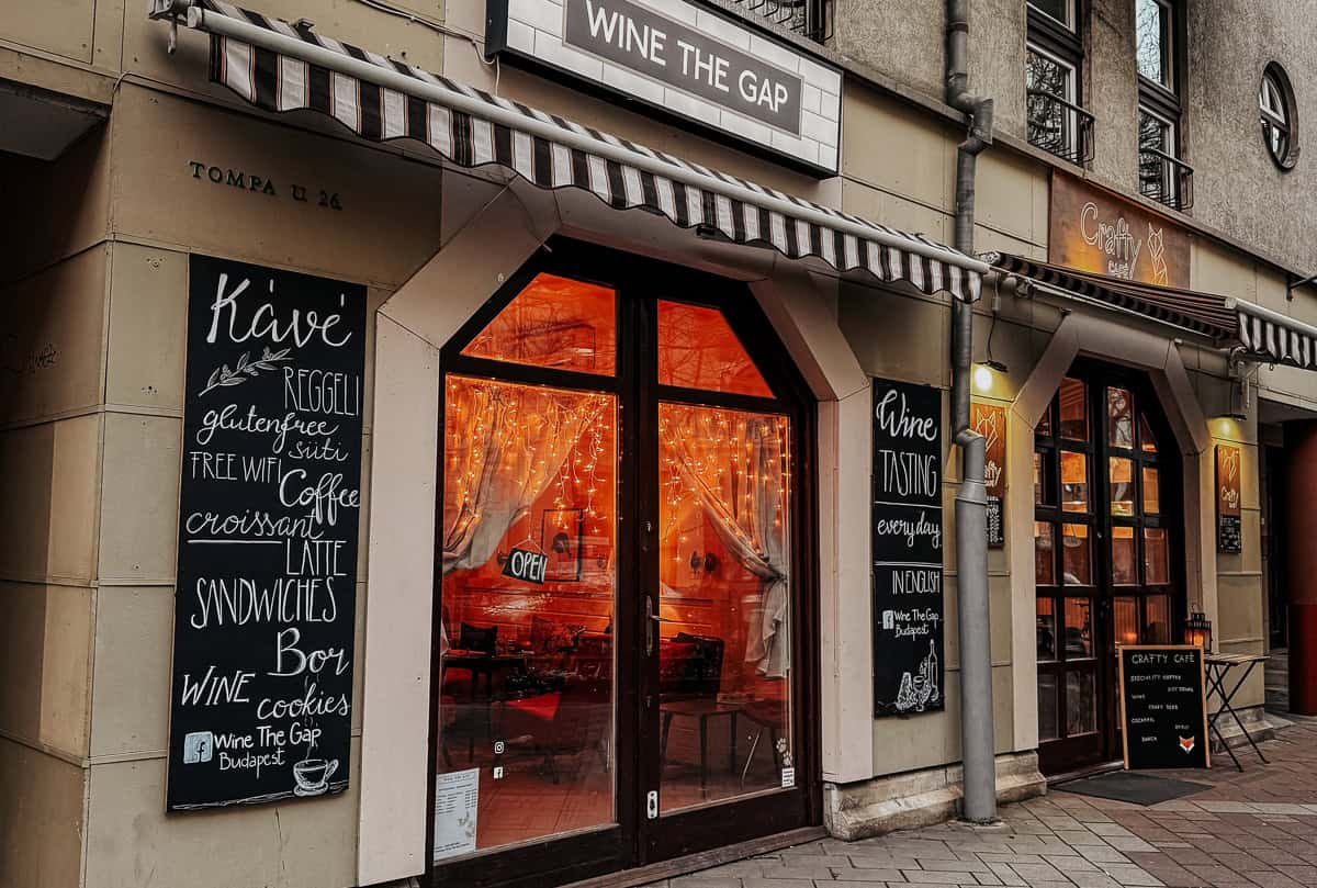 The exterior of "Wine The Gap," a wine bar and café with a striped awning and chalkboard signs advertising coffee, sandwiches, and wine tastings. The windows are adorned with twinkling lights, creating a welcoming atmosphere. ​​