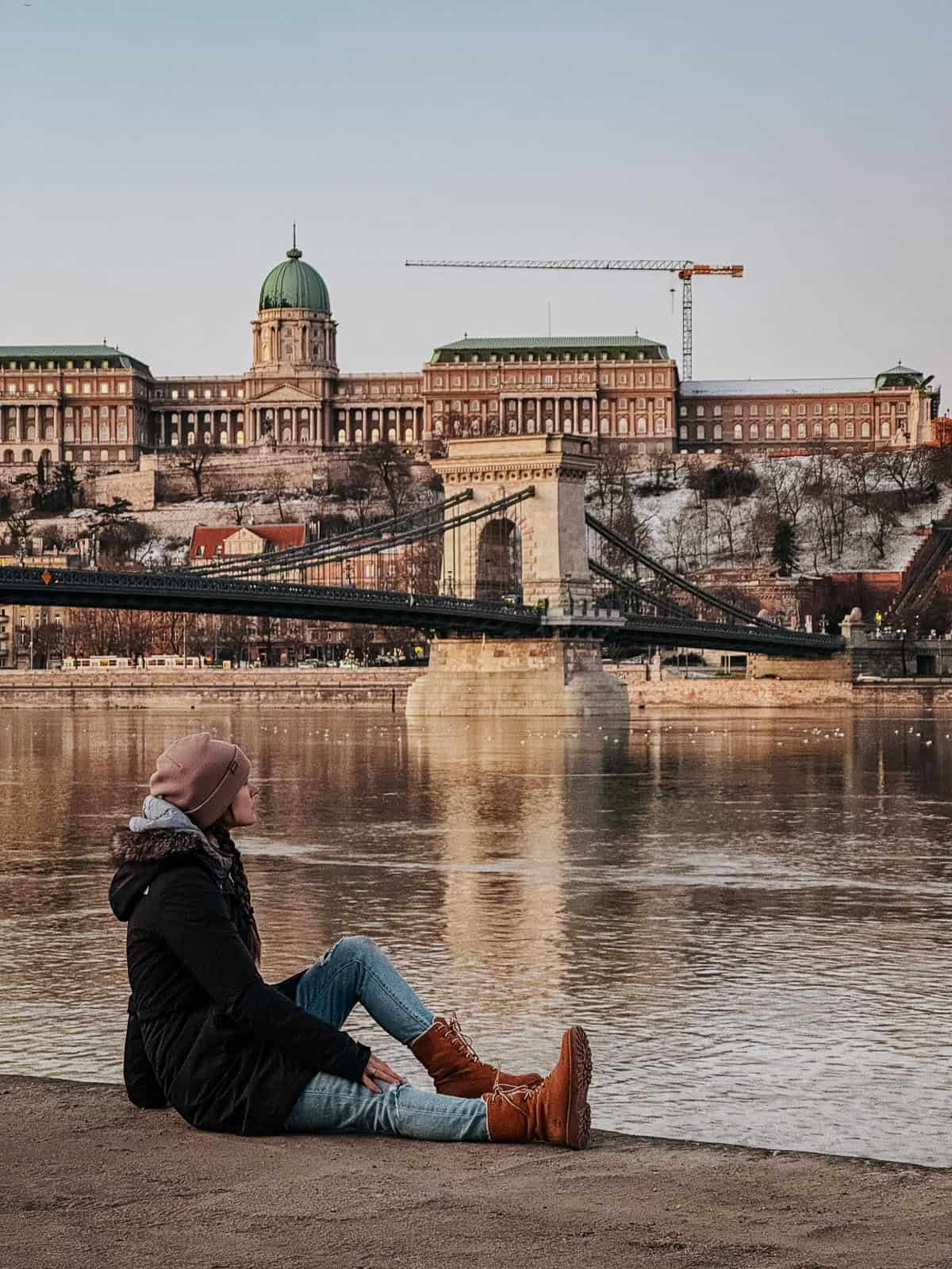 A person sits by the riverbank with Buda Castle in the background in Budapest, Hungary, under a clear sky.