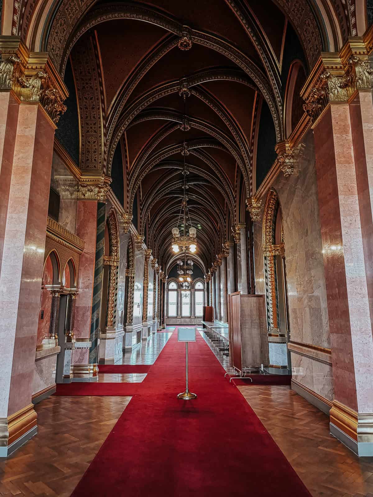 A grand hallway inside the Hungarian Parliament Building with a red carpet, high arches, and luxurious gold detailing on the walls and ceiling.